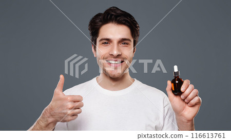 Masculine beauty. Handsome man approving face serum, showing thumb up gesture with small bottle, grey background Masculine beauty. Handsome man approving face serum, showing thumb up gesture with small bottle, grey background 116613761