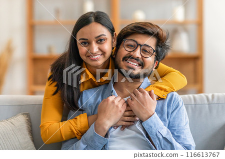 A happy Indian couple is seated on a comfortable sofa, smiling warmly at the camera. The woman is affectionately embracing her partner from behind, both appearing content and relaxed A happy Indian couple is seated on a comfortable sofa, smiling warmly at the camera. The woman is affectionately embracing her partner from behind, both appearing content and relaxed 116613767