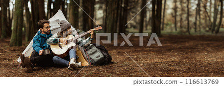 A couple sits by a tent in the middle of a forest. The woman plays an acoustic guitar as the man listens attentively. The campsite is surrounded by tall trees, panorama with copy space A couple sits by a tent in the middle of a forest. The woman plays an acoustic guitar as the man listens attentively. The campsite is surrounded by tall trees, panorama with copy space 116613769