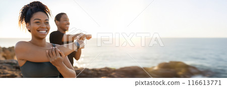 African American woman and man are stretching their arms in front of a beautiful sunset view over the ocean. They are standing on a rocky cliff, with the ocean stretching out behind them, copy space African American woman and man are stretching their arms in front of a beautiful sunset view over the ocean. They are standing on a rocky cliff, with the ocean stretching out behind them, copy space 116613771