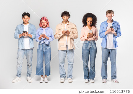 This image shows five diverse young adults, all standing in a line and looking down at their smartphones. They are dressed in casual clothing, and the background is a simple white wall. 116613776