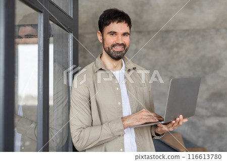 A man with short brown hair and a beard is standing in a modern office building. He is smiling and holding a laptop in his hands. 116613780