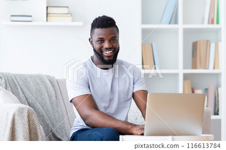 Black man with a beard sits on a couch in a home office, using a laptop computer. He is smiling and looks happy and relaxed. Room is decorated with white walls and a bookcase full of books. Black man with a beard sits on a couch in a home office, using a laptop computer. He is smiling and looks happy and relaxed. Room is decorated with white walls and a bookcase full of books. 116613784