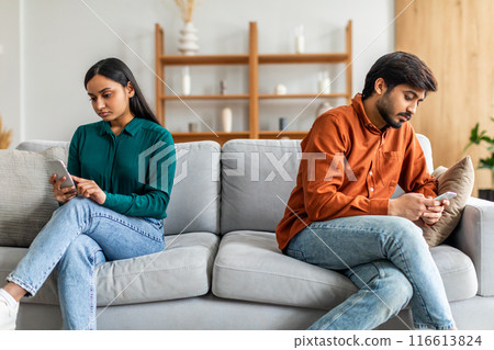 Indian couple sits on a gray couch in a modern living room, both absorbed in their smartphones. The room features light-colored walls, wooden shelves, and minimalistic decor 116613824