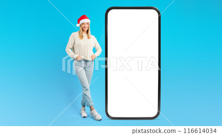 A woman wearing a Santa hat stands next to an oversized smartphone mockup with a white screen. She is casually dressed in jeans and sneakers, smiling on a vibrant blue background. A woman wearing a Santa hat stands next to an oversized smartphone mockup with a white screen. She is casually dressed in jeans and sneakers, smiling on a vibrant blue background. 116614034