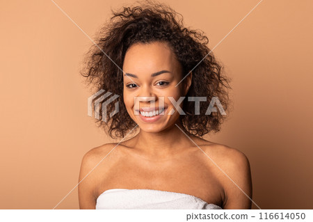 This image is a portrait of a young African American woman with dark curly hair smiling at the camera. She is wearing a white towel wrapped around her chest. The background is a solid beige color This image is a portrait of a young African American woman with dark curly hair smiling at the camera. She is wearing a white towel wrapped around her chest. The background is a solid beige color 116614050
