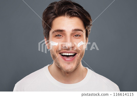 Male beauty and body care. Cheerful millennial man enjoying pampering cream on face, grey background 116614115