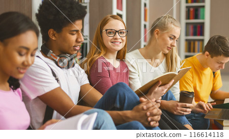 Concentrated international classmates sitting on wooden floor at library in campus and preparing for exams with books and laptops Concentrated international classmates sitting on wooden floor at library in campus and preparing for exams with books and laptops 116614118