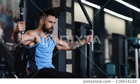 A man with tattoos works out on a cable machine in a gym. 116614120