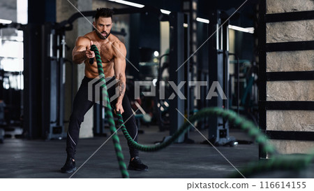 A muscular man is working out with battle ropes in a gym. He is wearing black shorts and shoes. He is focused on his workout. 116614155