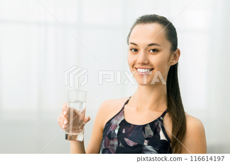 A woman in athletic wear holds a glass of water, smiling happily after a workout. 116614197