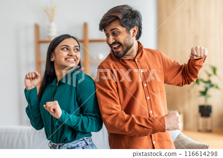 A young Indian couple joyfully dances together in their brightly lit living room, wearing casual clothes and expressing happiness through playful movements A young Indian couple joyfully dances together in their brightly lit living room, wearing casual clothes and expressing happiness through playful movements 116614298