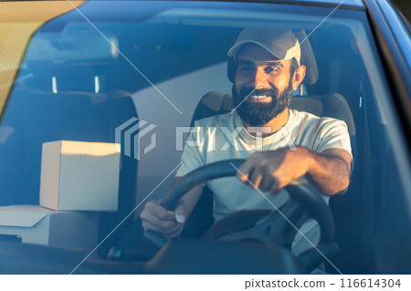 A cheerful Indian delivery driver navigates his vehicle through city streets early in the morning. He is seated behind the wheel, with packages visible beside him A cheerful Indian delivery driver navigates his vehicle through city streets early in the morning. He is seated behind the wheel, with packages visible beside him 116614304