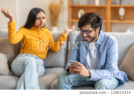 Indian man wearing glasses and a blue shirt is smiling and looking at his phone. Next to him, a woman in a yellow shirt is sitting with raised hands, displaying frustration Indian man wearing glasses and a blue shirt is smiling and looking at his phone. Next to him, a woman in a yellow shirt is sitting with raised hands, displaying frustration 116614314