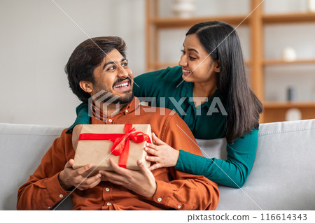 A smiling Indian couple enjoys a moment together as one presents a beautifully wrapped gift with a red ribbon to the other, in a warmly lit and cozy living room setting. 116614343