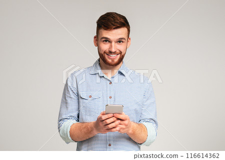 Young bearded man using his phone and smiling on studio background, websurfing or scrolling, copy space 116614362