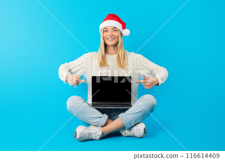 A woman wearing a Santa hat is seated on the floor, focused on using a laptop with blank display in front of her. She appears engaged and concentrated on the screen, surrounded by a cozy setting. 116614409