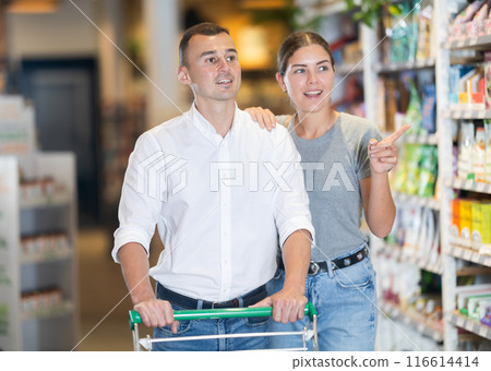 Married couple with trolley in trading floor browses eco product in bio shop search perfect goods Married couple with trolley in trading floor browses eco product in bio shop search perfect goods 116614414