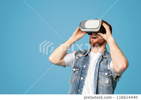 Excited young redhaired man enjoying virtual reality glasses experience, modern entertainment, blue studio background, free space 116614440