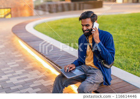 Indian man in a blue shirt and yellow t-shirt is sitting on a wooden bench in a park. He is engaged in conversation on his mobile phone while simultaneously working on his laptop Indian man in a blue shirt and yellow t-shirt is sitting on a wooden bench in a park. He is engaged in conversation on his mobile phone while simultaneously working on his laptop 116614446