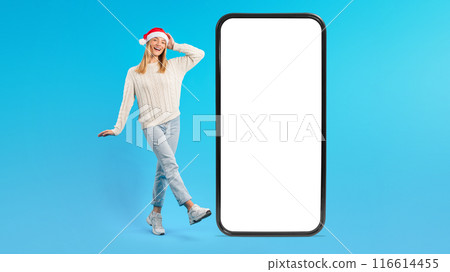 A cheerful young woman wearing a Santa hat and festive attire stands next to a large smartphone with white blank screen, posing playfully against a bright blue background. A cheerful young woman wearing a Santa hat and festive attire stands next to a large smartphone with white blank screen, posing playfully against a bright blue background. 116614455
