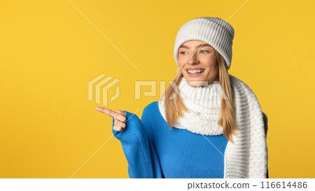A cheerful young woman dressed in a blue sweater, white scarf, and matching knit hat points to the side while standing against a vibrant yellow background, pointing at copy space A cheerful young woman dressed in a blue sweater, white scarf, and matching knit hat points to the side while standing against a vibrant yellow background, pointing at copy space 116614486