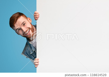 Cheerful young smiling guy looking out of white blank placard ready for text, information or product, blue background Cheerful young smiling guy looking out of white blank placard ready for text, information or product, blue background 116614520