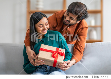 Indian man sitting on a couch is handing a gift to a woman next to him. The woman appears surprised and delighted as she receives the gift 116614535