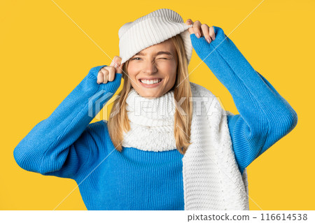 A woman is smiling cheerfully while adjusting her white winter hat. She is dressed in a blue sweater and matching white scarf, posing in front of a bright yellow background. 116614538