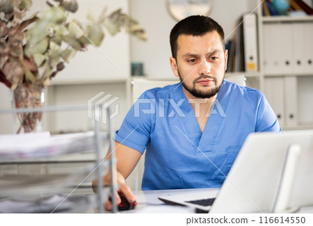 Man doctor sitting at table in office and using laptop Man doctor sitting at table in office and using laptop 116614550