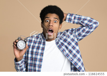 Late to college. Shocked guy holding alarm clock over light background Late to college. Shocked guy holding alarm clock over light background 116614555