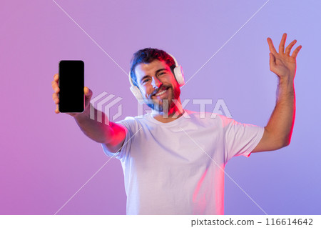 A man wearing headphones holds up a cell phone, with a focused expression on his face. He appears to be listening to music or talking on the phone. A man wearing headphones holds up a cell phone, with a focused expression on his face. He appears to be listening to music or talking on the phone. 116614642