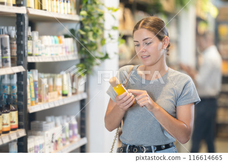 Positive young girl purchaser choosing cosmetic product in large supermarket 116614665