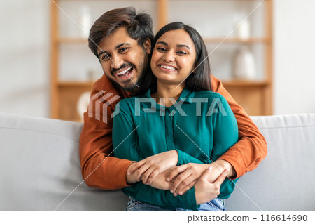 A smiling Indian couple is sitting on a comfortable sofa, warmly embracing each other in a well-lit living room. Both appear joyful and relaxed, with a calm and pleasant atmosphere in the background. A smiling Indian couple is sitting on a comfortable sofa, warmly embracing each other in a well-lit living room. Both appear joyful and relaxed, with a calm and pleasant atmosphere in the background. 116614690