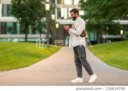 Indian man is walking down a sidewalk while engrossed in his cell phone, his attention focused on the screen rather than his surroundings, multitasking as he navigates the urban environment. 116614706