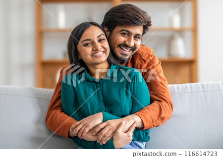 A happy Indian couple is sitting on a couch, with the man embracing the woman from behind. Both are smiling warmly, creating a heartwarming and affectionate atmosphere in a cozy living room. 116614723
