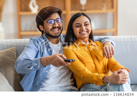 Indian man and woman are seated on a couch, holding a remote control. They appear to be watching television together, immersed in the content being displayed on the screen. Indian man and woman are seated on a couch, holding a remote control. They appear to be watching television together, immersed in the content being displayed on the screen. 116614834