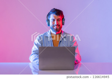 A man wearing headphones is sitting at a desk with a laptop in front of him. He appears to be enjoying music while working, with a smile on his face. The room is illuminated A man wearing headphones is sitting at a desk with a laptop in front of him. He appears to be enjoying music while working, with a smile on his face. The room is illuminated 116614945