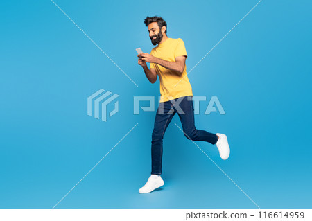 An adult Indian man wearing a yellow shirt is jogging outdoors with his attention focused on a smartphone screen. 116614959