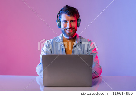 A man is seated in front of a laptop, wearing headphones and engaging with the screen. He appears focused and attentive, possibly working or listening to music. A man is seated in front of a laptop, wearing headphones and engaging with the screen. He appears focused and attentive, possibly working or listening to music. 116614990