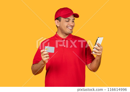 A cheerful delivery worker in a red uniform and cap is seen holding a credit card and a smartphone. He appears engaged as he looks at the smartphone screen, standing against a vivid yellow background. 116614996