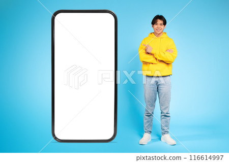 Asian young guy dressed in a yellow hoodie and blue jeans stands with arms crossed next to a large blank smartphone screen. The background is bright blue, creating a clean and modern look. 116614997