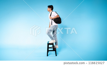 Asian guy is standing on a stool while wearing a backpack. He appears to be preparing for a task or adventure, showcasing a sense of readiness and determination in his posture. Asian guy is standing on a stool while wearing a backpack. He appears to be preparing for a task or adventure, showcasing a sense of readiness and determination in his posture. 116615027