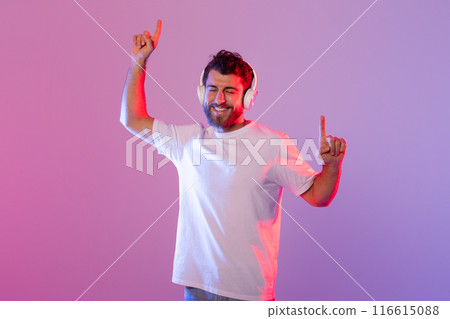 A cheerful man wearing wireless headphones is dancing and enjoying music. The background features vibrant, colorful studio lighting, enhancing the lively atmosphere. A cheerful man wearing wireless headphones is dancing and enjoying music. The background features vibrant, colorful studio lighting, enhancing the lively atmosphere. 116615088