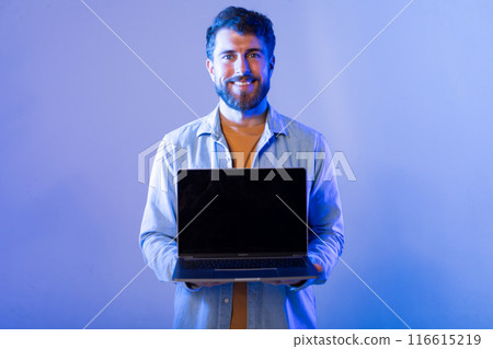 A smiling man wearing a light jacket stands indoors against a blue background, holding a laptop with black screen with both hands as he presents it toward the camera. 116615219