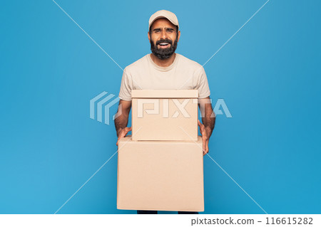 Indian delivery man dressed in a beige t-shirt and cap holds two cardboard boxes. He stands against a bright blue background, smiling. 116615282