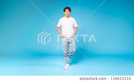 Asian guy in a white t-shirt is standing upright in front of a plain blue background. His posture is relaxed, with arms by his side and feet grounded on the floor. 116615310
