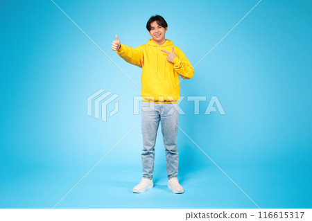 Asian guy wearing a bright yellow hoodie is enthusiastically giving a thumbs up gesture. He appears cheerful and positive in his body language, showing approval or agreement. Asian guy wearing a bright yellow hoodie is enthusiastically giving a thumbs up gesture. He appears cheerful and positive in his body language, showing approval or agreement. 116615317