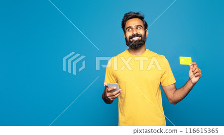 Indian man wearing a yellow shirt is seen holding a yellow card in one hand and a cell phone in the other, copy space 116615365