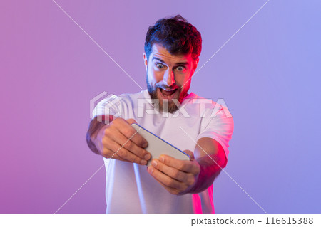 A man with a beard and wearing a white shirt is enthusiastically playing a game on his smartphone. The room is lit with vibrant pink and purple lighting, indicating it is nighttime. 116615388